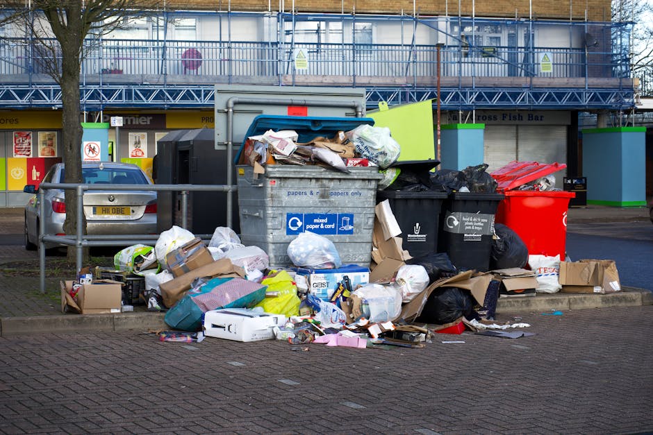 A collection of overflowing rubbish bins and scattered waste on a paved parking area in front of a commercial building. The heap includes a large grey mixed paper and card bin with its lid open, revea