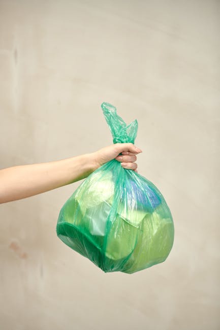 A pile of mixed household rubbish, including flattened cardboard boxes with visible printed labels, crumpled brown paper bags, and a large white fabric or sack, all gathered against an external brick 