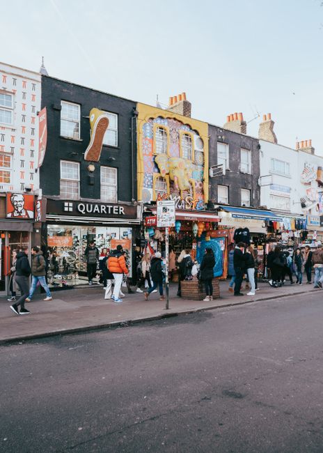 A busy urban street scene in Camden High Street featuring a mix of commercial buildings and retail shops. In the foreground, a red double-decker bus and a small red car are traveling along the asphalt