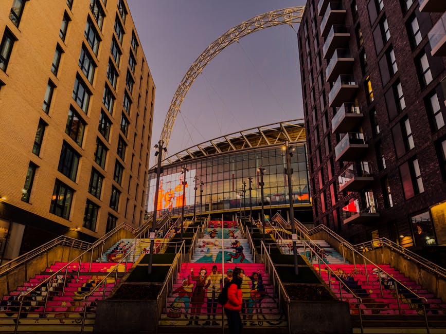 A modern, large-scale sports stadium named Emirates Stadium is depicted with a curved roof extending over the facade, which is composed of glass panels and structural metal elements. In the foreground